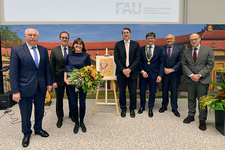 Umringt von den Honoratioren steht die Preisträgerin mit einem Blumenstrauß in der Hand neben einer Staffelei, auf der ein Bild mit dem Porträt von Jakob Herz zu sehen ist und lächelt in die Kamera.